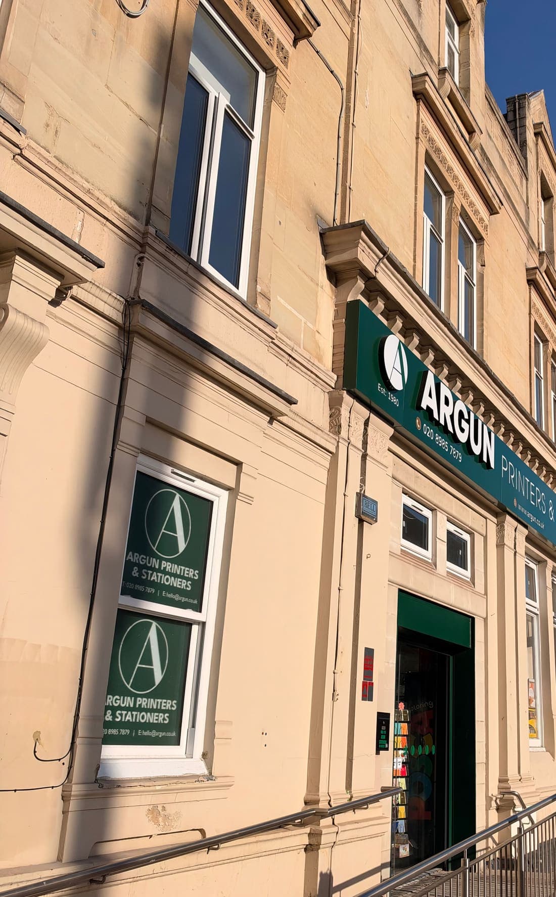 The front of 20 Amhurst Road, a pale stone building with the green Argun Printers and Stationers sign above the door and a ramp with handrails up to the entrance.