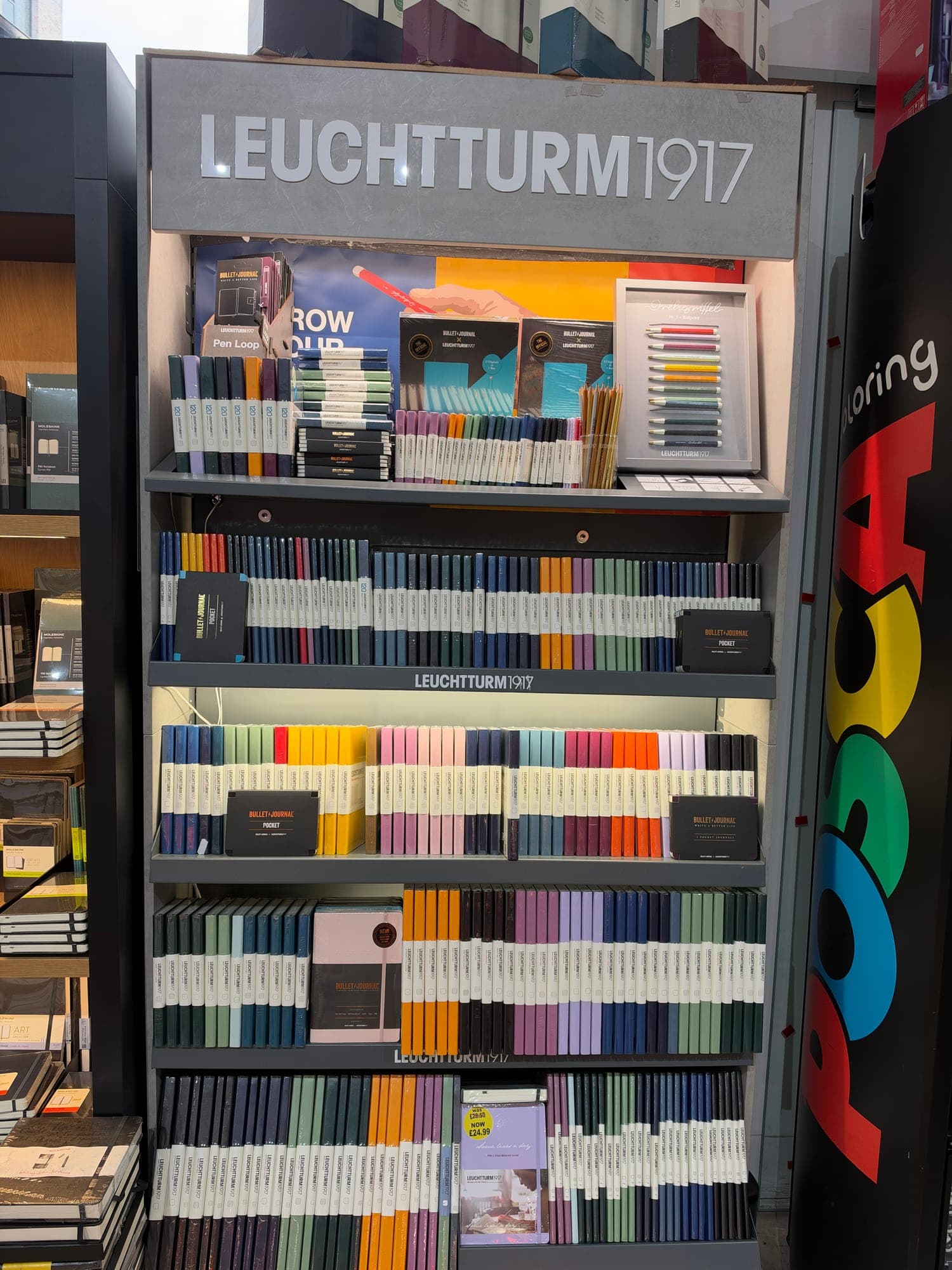 A lit Leuchtturm1917 wall of notebooks and bullet journals in rows of colours, next to a Posca marker display.